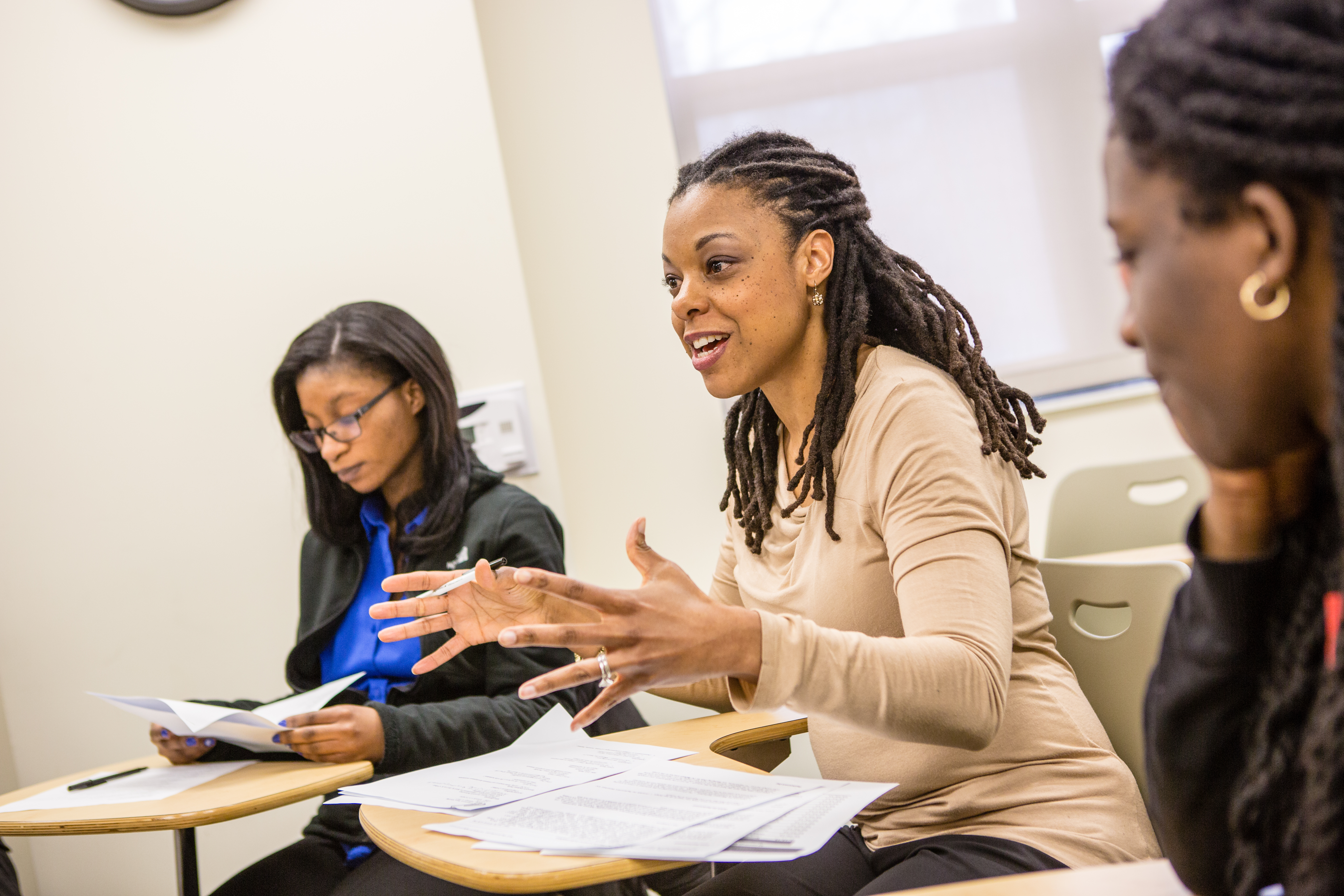 A female student sits at their desk in the classroom with two other students to their right and left. They are speaking in class with their hands mid-gesture.