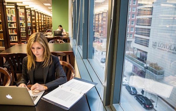 A woman sits at a table studying from library books in a high-rise skyscraper with a large window overlooking the city