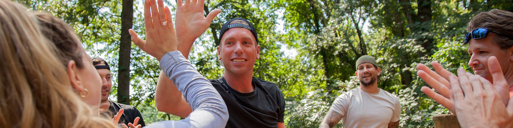 A group of people outdoors in a forested area are high-fiving each other, appearing upbeat and smiling. One person in the center in a black shirt and cap looks directly at the camera while engaged in the high-five. The atmosphere is lively and energetic.