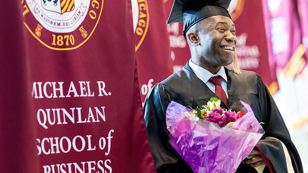 Person in graduation hat and gown smiling and holding flowers next to a maroon banner that says Person in graduation hat and gown smiling and holding flowers next to a maroon banner that says