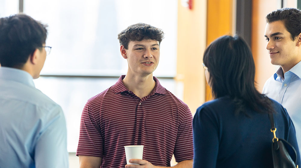 A student speaks with a group other students, alumni, and faculty A student speaks with a group other students, alumni, and faculty