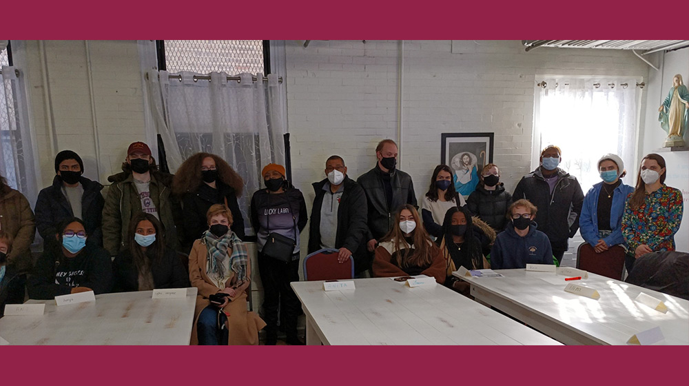 A large group of people gather for a group photo. Some are seated at square white tables while others stand in a row behind them. All wear PPE masks