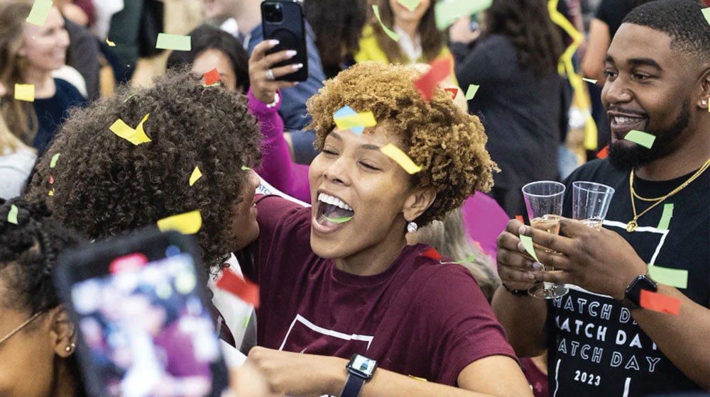 Loyola Chicago celebration with two women hugging in a crowd with confetti falling from the ceiling. A man in the background smiling, holding a drink.