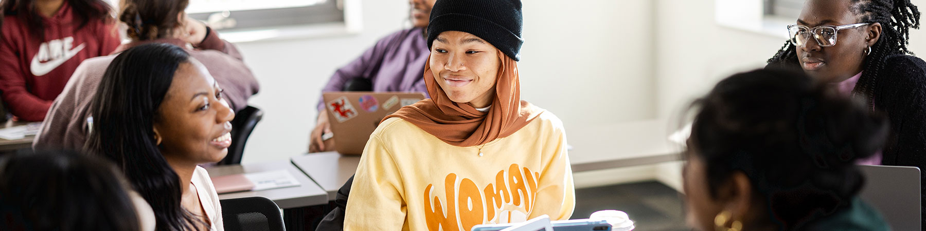 A diverse group of Loyola University Chicago students interact with each other, smiling, in a classroom. There is one group in focus, with others blurred in the background, all with laptop computers open on their desks.