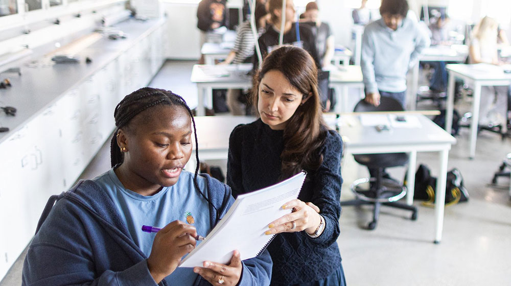 An instructor reviews work with a student in a classroom with other students woking at tables in the background. An instructor reviews work with a student in a classroom with other students woking at tables in the background.